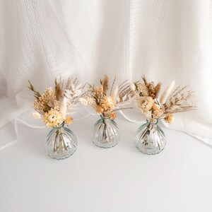 May include: Three small, clear glass vases with a ribbed design, each holding a small arrangement of dried flowers in shades of beige, cream, and light yellow. The vases sit on a white surface against a white, textured backdrop.