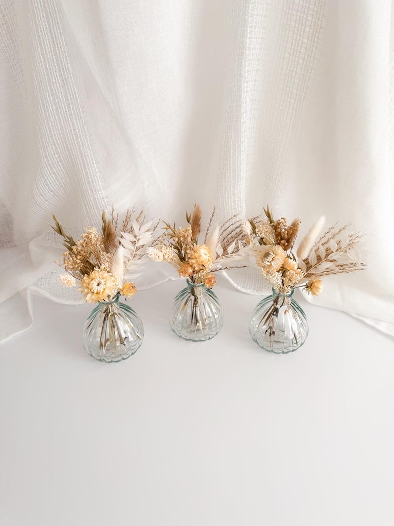 May include: Three small, clear glass vases with a ribbed design, each holding a small arrangement of dried flowers in shades of beige, cream, and light yellow. The vases sit on a white surface against a white, textured backdrop.