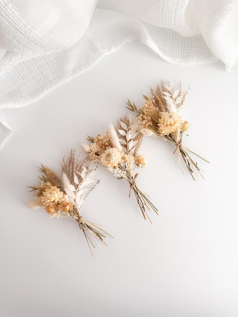 May include: Three small bouquets of dried flowers in shades of beige, cream, and tan. Each bouquet is tied with twine and features a variety of textures, including feathery grasses and delicate blooms. The background is a soft white.