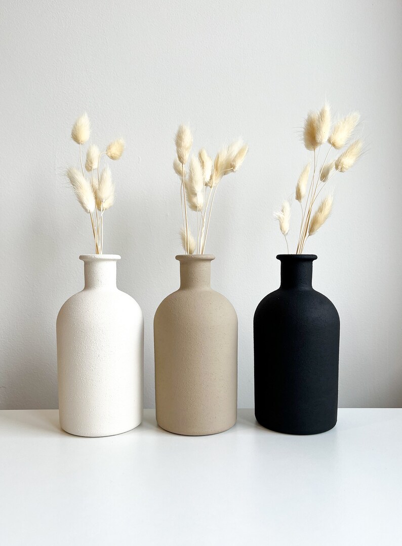 Pode incluir: Tr&ecirc;s vasos de cer&acirc;mica em branco, bege e preto com grama de pampas seca em cada um.