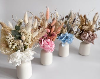 May include: Four small, white ceramic vases each hold a unique arrangement of dried flowers. The bouquets feature a variety of colors, including white, pink, blue, and beige, creating a natural and elegant aesthetic. The vases are arranged in a row.