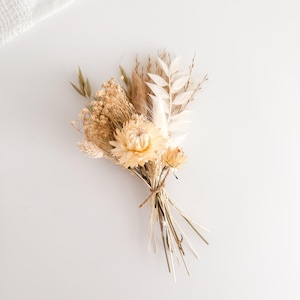 May include: A small, dried flower bouquet featuring a variety of beige and cream-colored blooms and grasses. The bouquet is tied with twine and rests on a white surface. The arrangement includes a large, light yellow flower as the focal point.