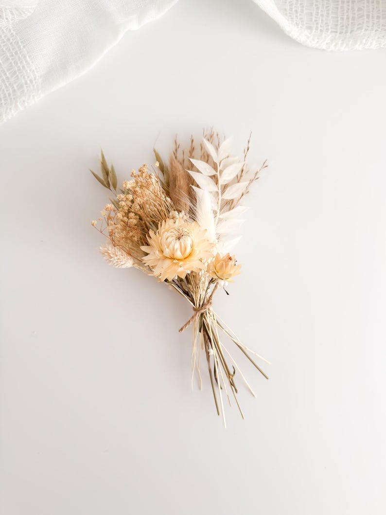 May include: A small, dried flower bouquet featuring a variety of beige and cream-colored blooms and grasses. The bouquet is tied with twine and rests on a white surface. The arrangement includes a large, light yellow flower as the focal point.