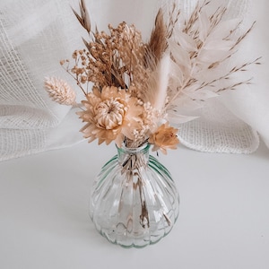 May include: A small, clear glass vase with a ribbed design holds a bouquet of dried flowers. The arrangement includes beige and tan blooms, feathery grasses, and delicate stems. The vase sits on a white surface, with a white fabric backdrop.