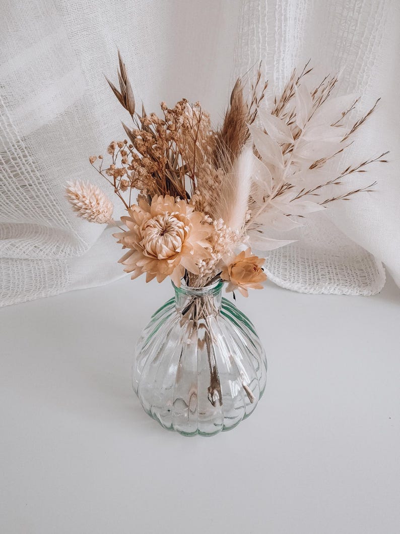 May include: A small, clear glass vase with a ribbed design holds a bouquet of dried flowers. The arrangement includes beige and tan blooms, feathery grasses, and delicate stems. The vase sits on a white surface, with a white fabric backdrop.