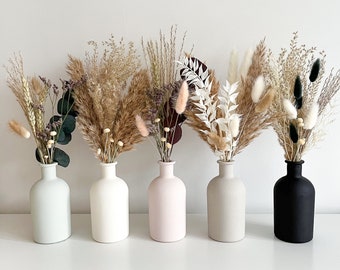 May include: Five small vases in different colors, each filled with dried pampas grass and other dried flowers. The vases are arranged in a row on a white surface.