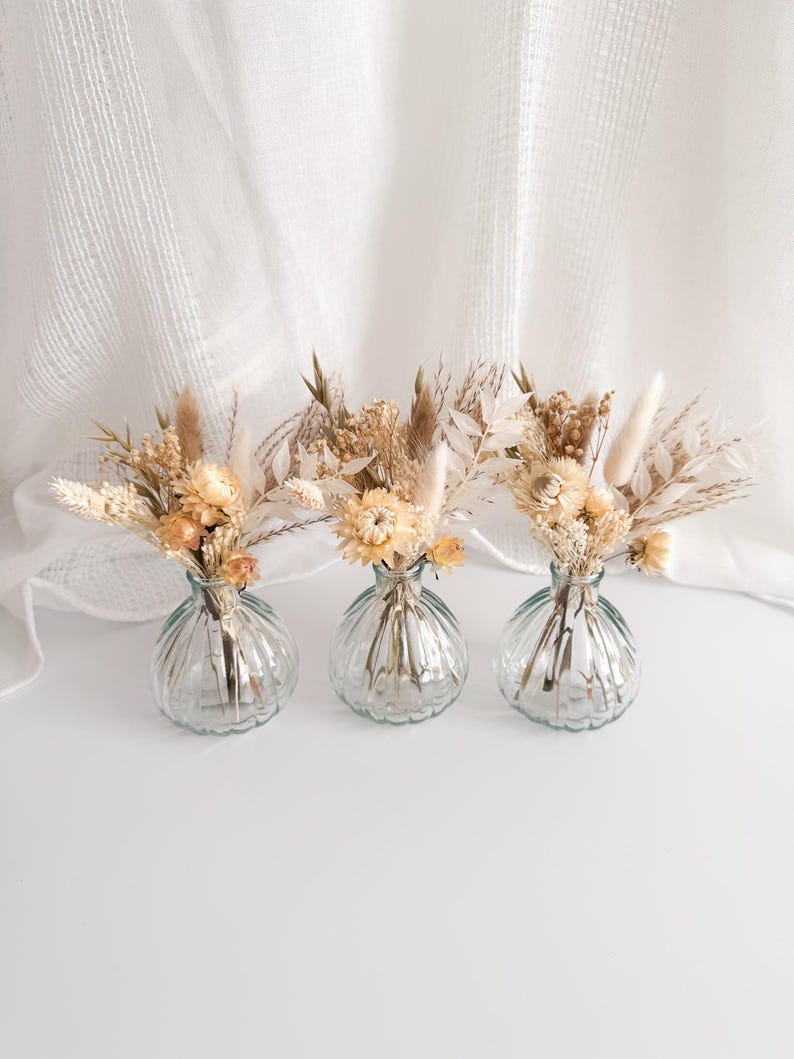 May include: Three small, clear glass vases with a ribbed design, each holding a bouquet of dried flowers in shades of beige, cream, and tan. The vases sit on a white surface against a white, textured backdrop. The floral arrangements include various grasses and delicate blooms.
