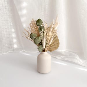 May include: A small, cream vase displays a dried floral arrangement. The arrangement features green eucalyptus leaves, beige pampas grass, and a dried palm leaf. The vase is on a white surface, with a white curtain in the background.