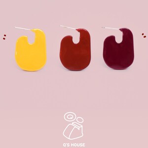 Peut inclure: Trois boucles d'oreilles de forme abstraite, jaune, rouge et bordeaux, chacune avec une tige argentée. Les boucles d'oreilles sont présentées sur un fond rose clair. Le logo de la marque "Q's HOUSE" est visible en bas de l'image.