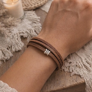 May include: A multi-strand brown suede leather bracelet featuring a small, silver-toned metal charm shaped like a butterfly. The bracelet wraps around the wrist in three distinct layers, showcasing a textured finish on the leather cords.