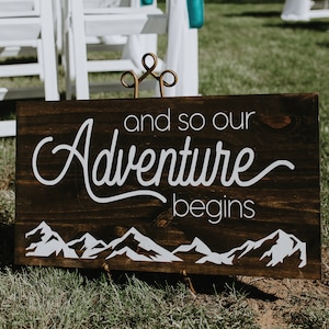 May include: A wooden sign with white text that reads "and so our Adventure begins" with a mountain range graphic at the bottom. The sign is propped up on a metal stand and is sitting on green grass.