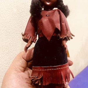May include: A vintage doll with dark skin, wearing a burgundy leather-like outfit with fringe detailing. The doll has black hair, a matching headband, and burgundy boots. The doll's skirt is black.
