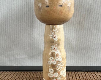 Puede incluir: Una muñeca japonesa de madera kokeshi con cabeza redonda y cara pintada. La muñeca lleva un kimono blanco y dorado con un patrón floral.