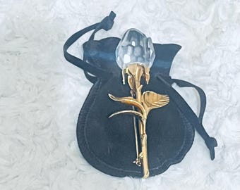 May include: A crystal rose brooch with a gold-tone stem and leaves, resting on a black velvet pouch. The rose has a faceted, clear crystal bloom. The brooch is a decorative accessory.
