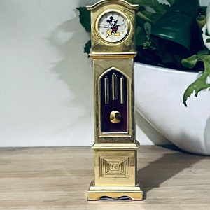 May include: A gold-colored Mickey Mouse grandfather clock with a white face and black hands. The clock has a pendulum and chimes.