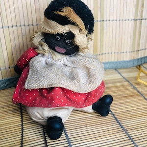 May include: A handmade rag doll with black fabric for the head and limbs, a brown fabric head wrap, and a red and white polka dot dress. The doll is sitting on a woven bamboo mat.