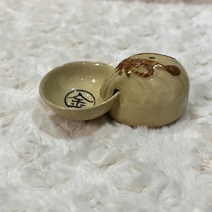 Puede incluir: Un pequeño cuenco de cerámica beige con un diseño marrón y una tapa a juego. El cuenco tiene un diseño circular con el carácter chino para "oro" en negro.