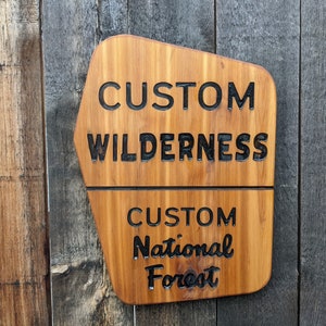 May include: A wooden sign with a rustic, weathered look. The sign is shaped like a mountain peak and has the text "CUSTOM WILDERNESS CUSTOM National Forest" in black lettering.