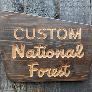 May include: A rustic wooden sign with the text "CUSTOM NATIONAL FOREST" carved into the surface. The sign is dark brown with a light brown wood grain.