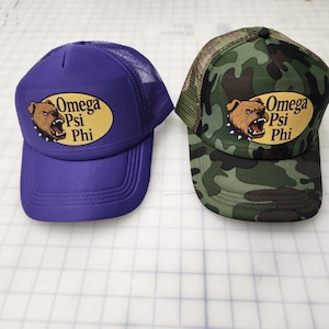 May include: Two baseball caps. One is purple with a mesh back, the other is camouflage. Both feature the text "Omega Psi Phi" and a bulldog graphic on a yellow oval.