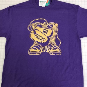 Puede incluir: Camiseta morada con un gráfico dorado que presenta placas de identificación militares y botas de combate. Las placas de identificación dicen "MILITARY BRUHZ". También se incluye un diseño de collar de cuentas. La camiseta tiene cuello redondo y mangas cortas.