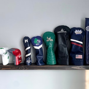 May include: A row of eight golf club head covers on a dark brown wooden shelf. The covers are in various colors and designs, including green, blue, red, white, and black. Some of the covers have embroidered logos or text.