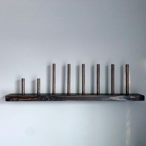 May include: A dark brown wooden shelf with nine wooden dowels of varying heights. The shelf is mounted on a white wall.