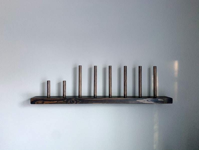 May include: A dark brown wooden shelf with nine wooden dowels of varying heights. The shelf is mounted on a white wall.