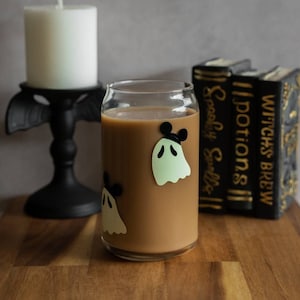 May include: A glass with a brown beverage and ghost stickers with Mickey Mouse ears. The glass is on a wooden surface with a white candle and a stack of books with the titles "Witch's Brew", "Potions", and "Spooky Spells" visible in the background.