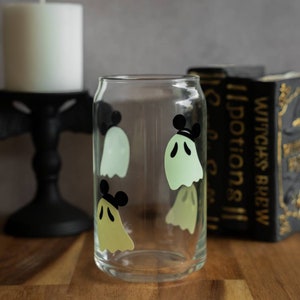 May include: A clear glass with a design of four green ghosts with black Mickey Mouse ears. The glass is sitting on a wooden surface.