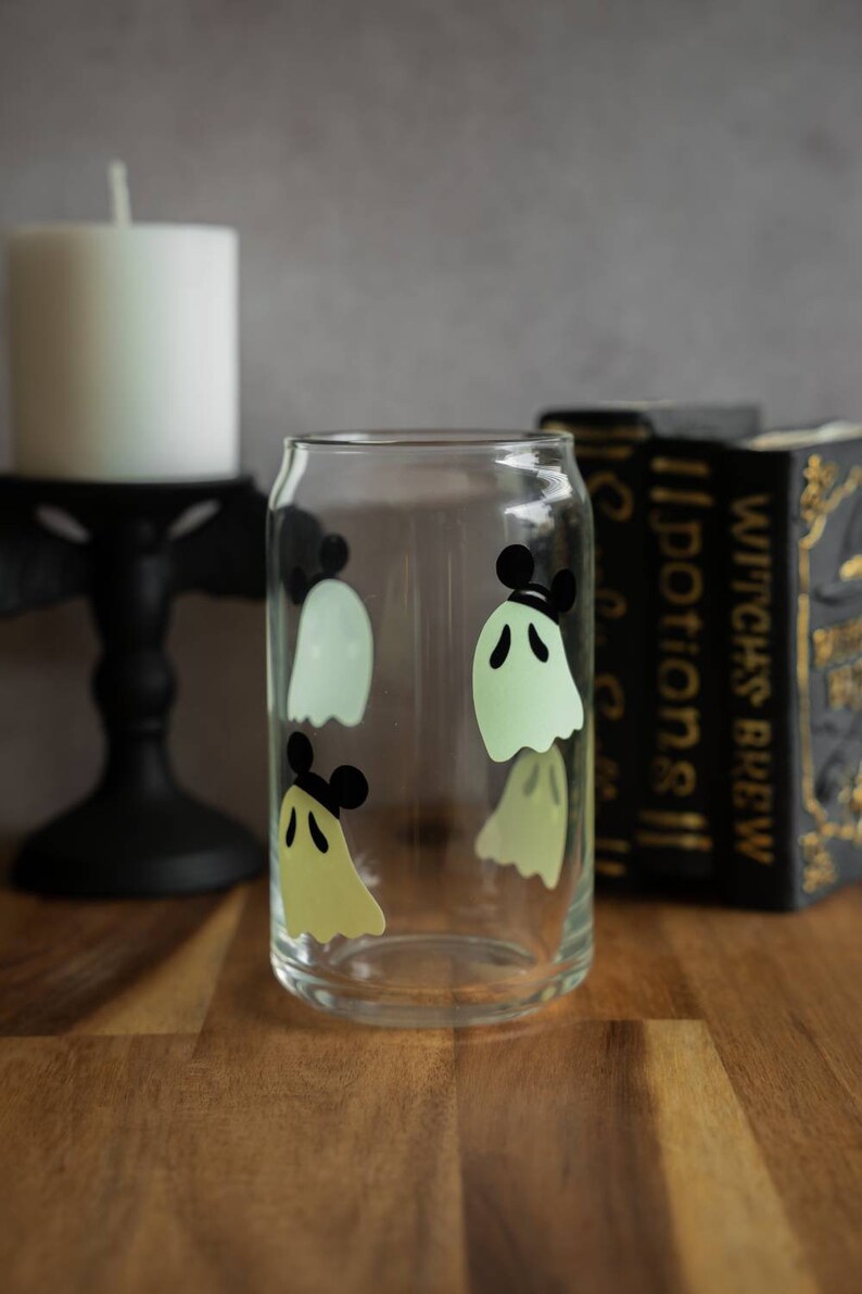 May include: A clear glass with a design of four green ghosts with black Mickey Mouse ears. The glass is sitting on a wooden surface.