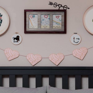 May include: A pink heart garland with a white and pink patterned design hangs on a wall above a black headboard. Two small embroidery hoops with the names "Buddy" and "Brides" are on the wall. Two white plates with cactus designs are on the wall.