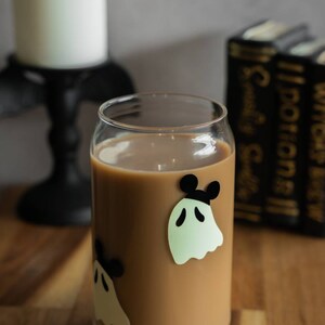 May include: A glass with a brown beverage and ghost stickers with black Mickey Mouse ears. The glass is on a wooden surface.