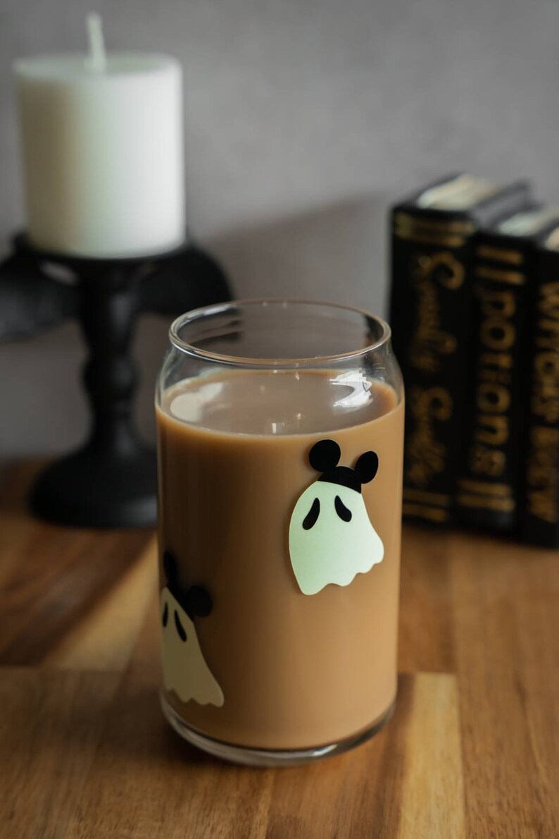 May include: A glass with a brown beverage and ghost stickers with black Mickey Mouse ears. The glass is on a wooden surface.