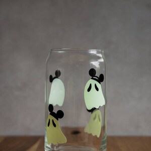 May include: A clear glass with a design of four ghosts with Mickey Mouse ears. The ghosts are in shades of green and yellow.