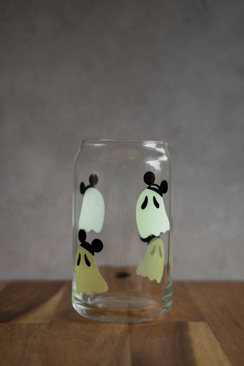 May include: A clear glass with a design of four ghosts with Mickey Mouse ears. The ghosts are in shades of green and yellow.