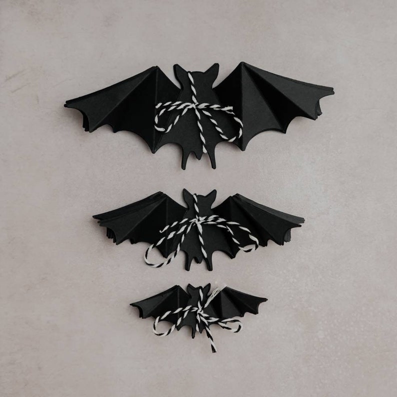 May include: Three black paper bats of different sizes, each tied with white string, are arranged in a descending order on a light gray background.