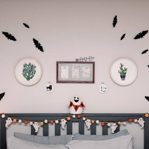May include: A gray headboard with a white and orange garland of ghosts and pom poms. There are black bats and two white plates with cactus illustrations on the wall behind the headboard. A white ghost with the word "BOO" on it is sitting on the headboard.
