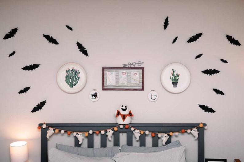 May include: A gray headboard with a white and orange garland of ghosts and pom poms. There are black bats and two white plates with cactus illustrations on the wall behind the headboard. A white ghost with the word "BOO" on it is sitting on the headboard.
