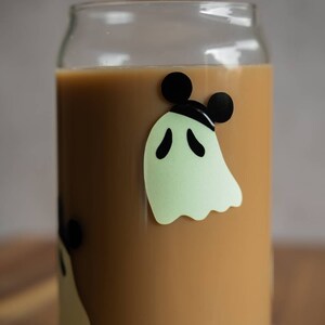 May include: A glass can with a brown liquid inside. The can has a ghost with Mickey Mouse ears on it. The ghost is green and the ears are black.