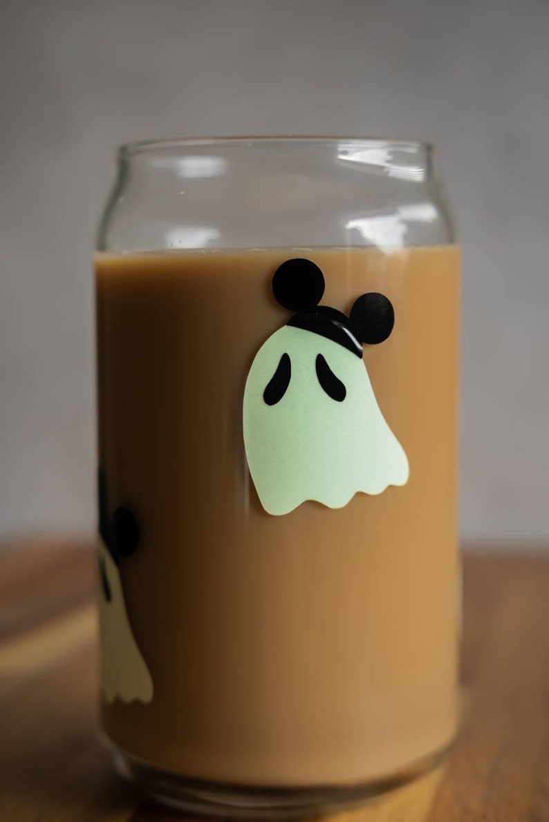 May include: A glass can with a brown liquid inside. The can has a ghost with Mickey Mouse ears on it. The ghost is green and the ears are black.