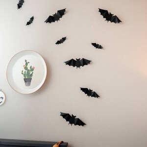 May include: A white wall decorated with black paper bats. A white plate with a green cactus illustration hangs on the wall. A black bed frame with a garland of orange and white pom-poms is at the bottom of the image.