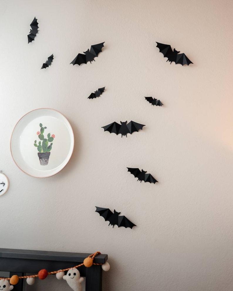 May include: A white wall decorated with black paper bats. A white plate with a green cactus illustration hangs on the wall. A black bed frame with a garland of orange and white pom-poms is at the bottom of the image.