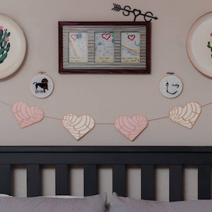May include: A pink and white heart-shaped garland hangs above a black headboard with a white and grey patterned pillow. Two plates with cactus designs are on the wall, along with a framed picture with three smaller pictures inside.