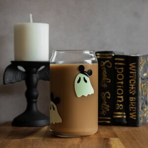 May include: A glass with a brown beverage and ghost stickers with Mickey Mouse ears. The glass is sitting on a wooden table next to a white candle and a stack of books with the titles "Witch's Brew", "Potions", and "Spooky Spells".