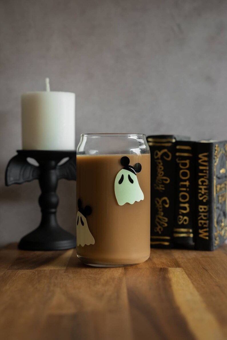 May include: A glass with a brown beverage and ghost stickers with Mickey Mouse ears. The glass is sitting on a wooden table next to a white candle and a stack of books with the titles "Witch's Brew", "Potions", and "Spooky Spells".