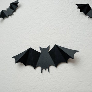 May include: Three black paper bats are hanging on a white wall. The bats are cut out in a simple silhouette shape.