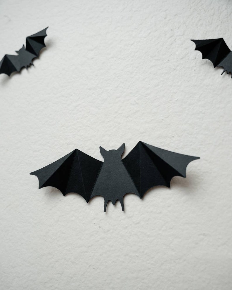 May include: Three black paper bats are hanging on a white wall. The bats are cut out in a simple silhouette shape.