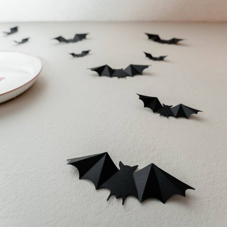 May include: Black paper bats are scattered on a white surface. The bats are cut out in a simple shape and are all facing the same direction.