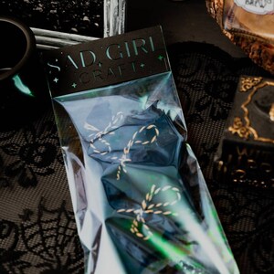 May include: A clear plastic bag with a black card that says "Sad Girl Craft" and contains two gold and black twine bows.